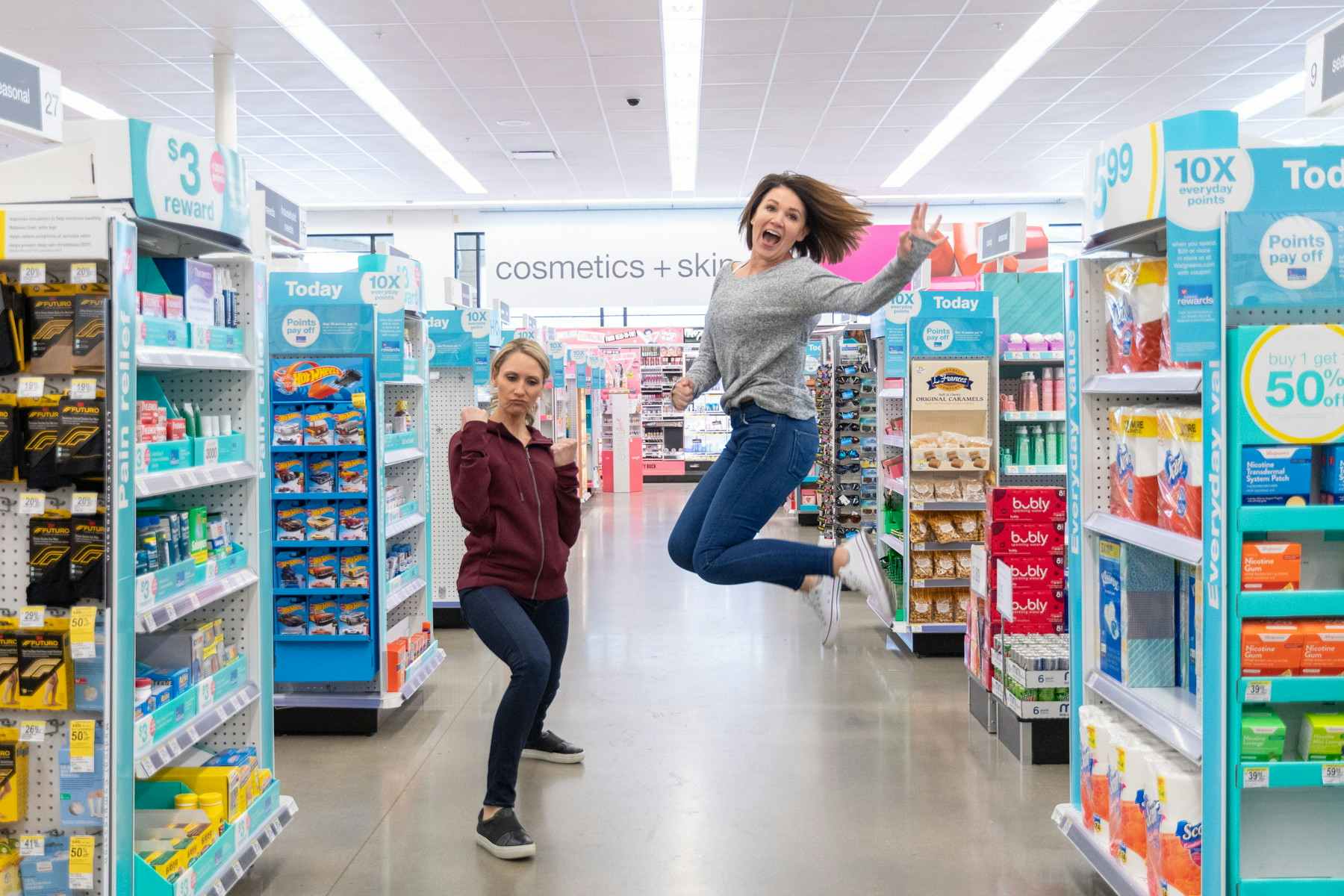 Two women inside Walgreens, one of them posing and the other one jumping into the air.