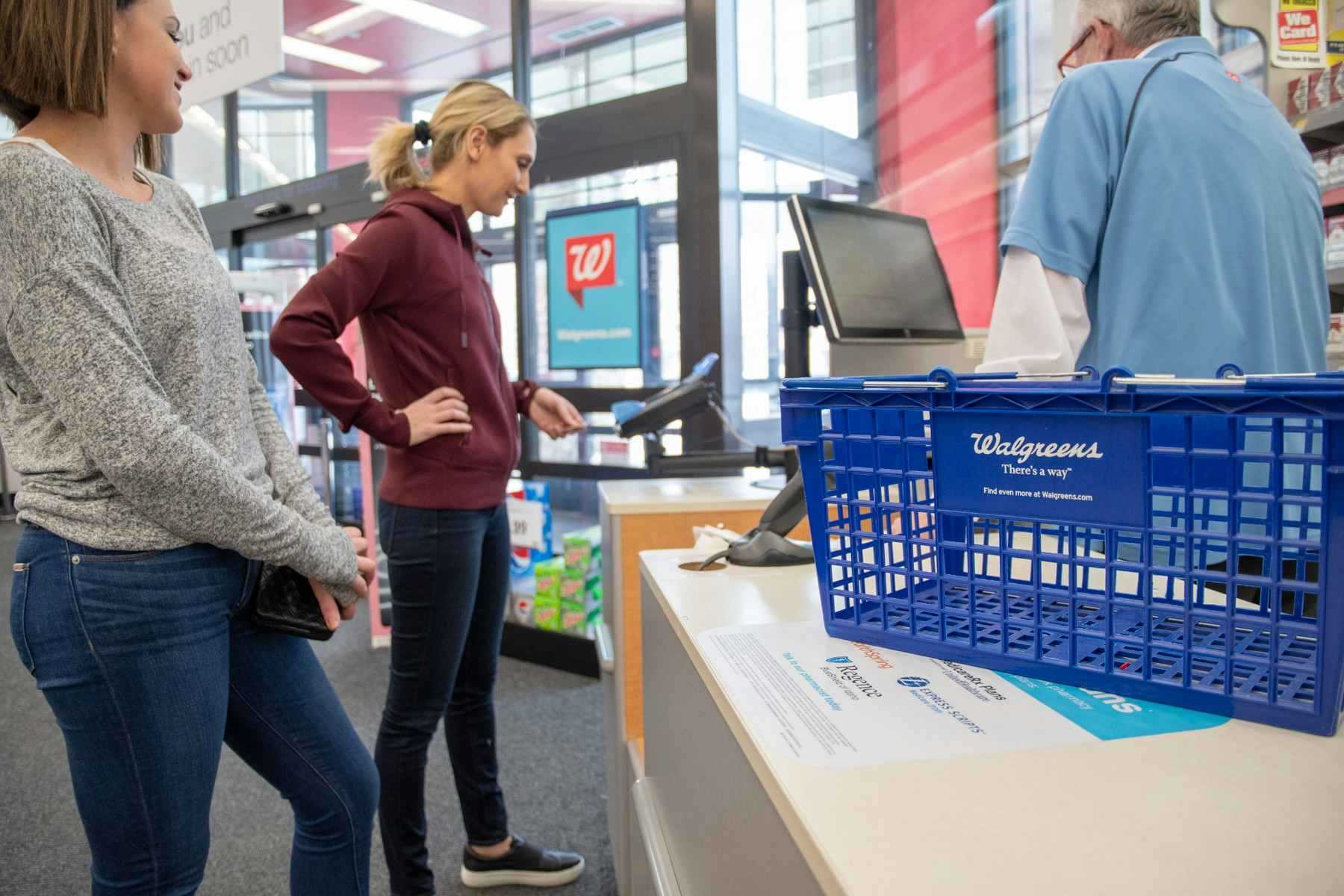 A Walgreens shopping basket on the counter. Two women standing at the checkout with one using the credit card reader to purchase items. 