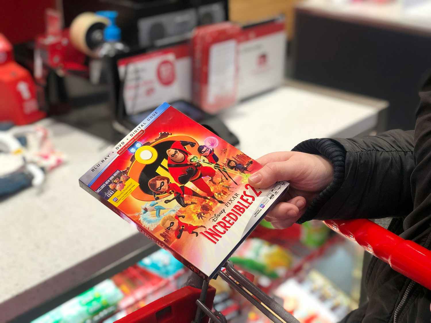 A person's hand holding a DVD case for Incredibles 2 above a Target shopping cart at the return counter at Target.