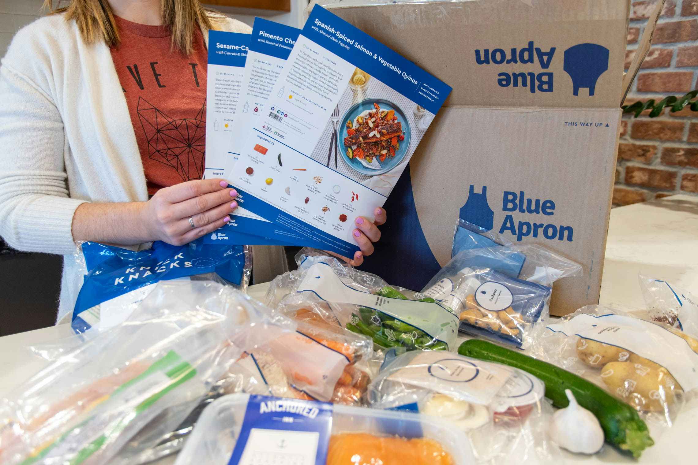 A woman holding the Blue Apron meal kit recipe cards next to the meal kit box and food.