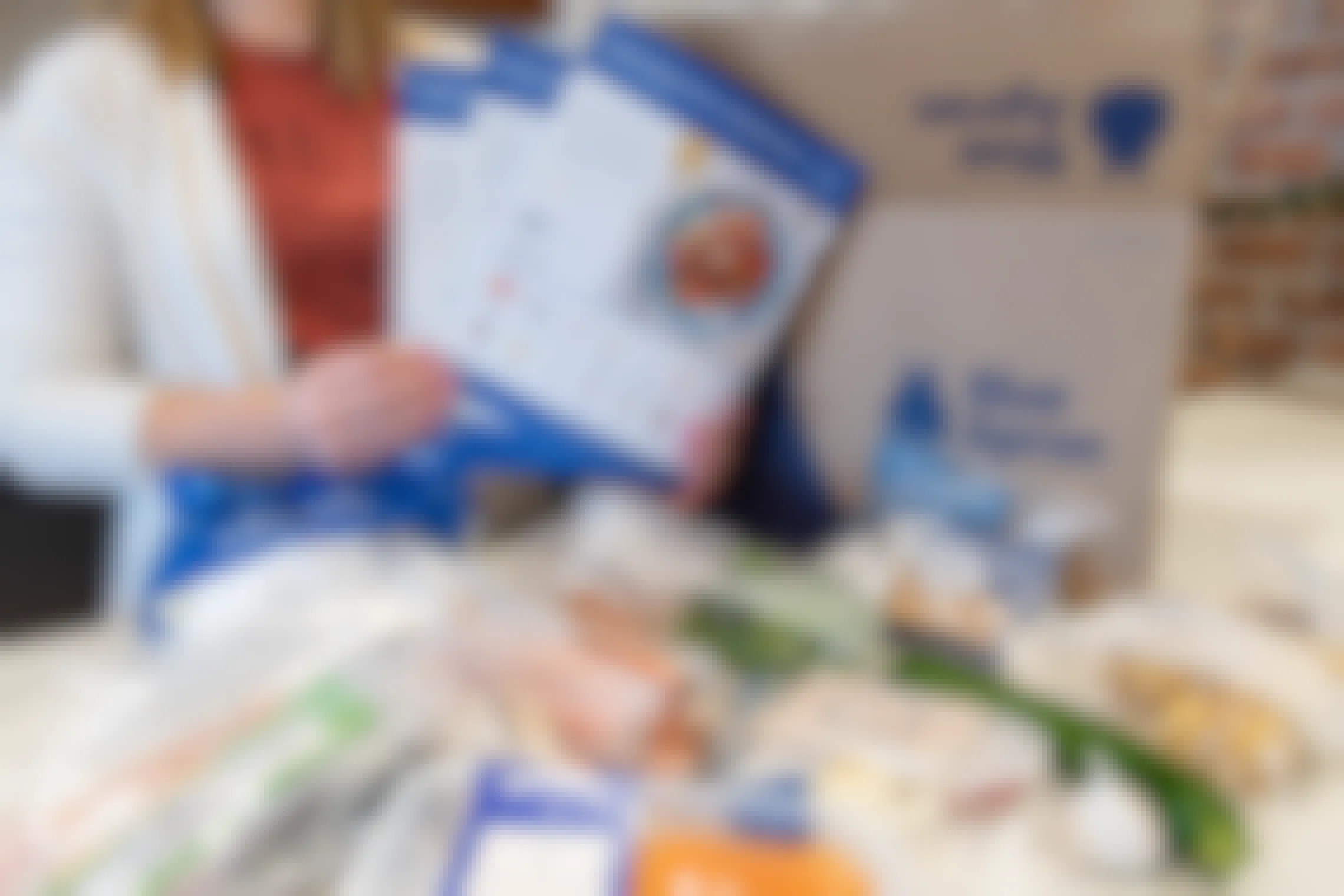 A woman holding the Blue Apron meal kit recipe cards next to the meal kit box and food.