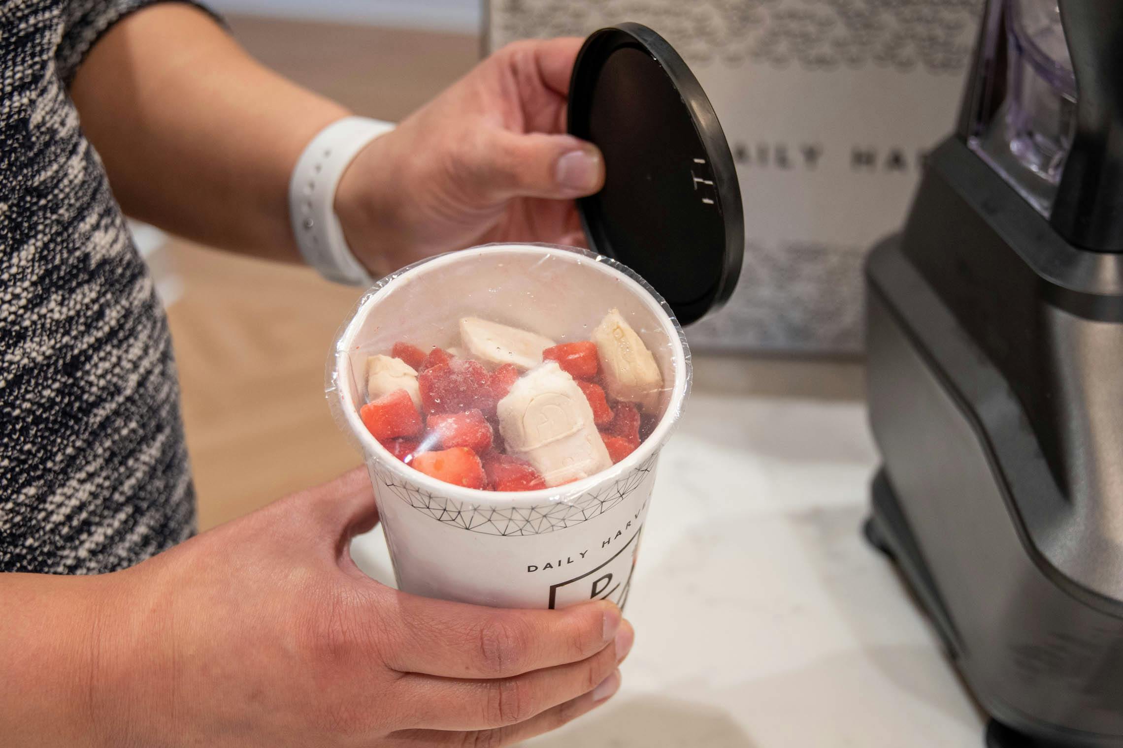 The inside of a smoothie cup showing strawberries and other fruit in front of a A woman pulling out the cups of food from a Daily Harvest meal kit box.