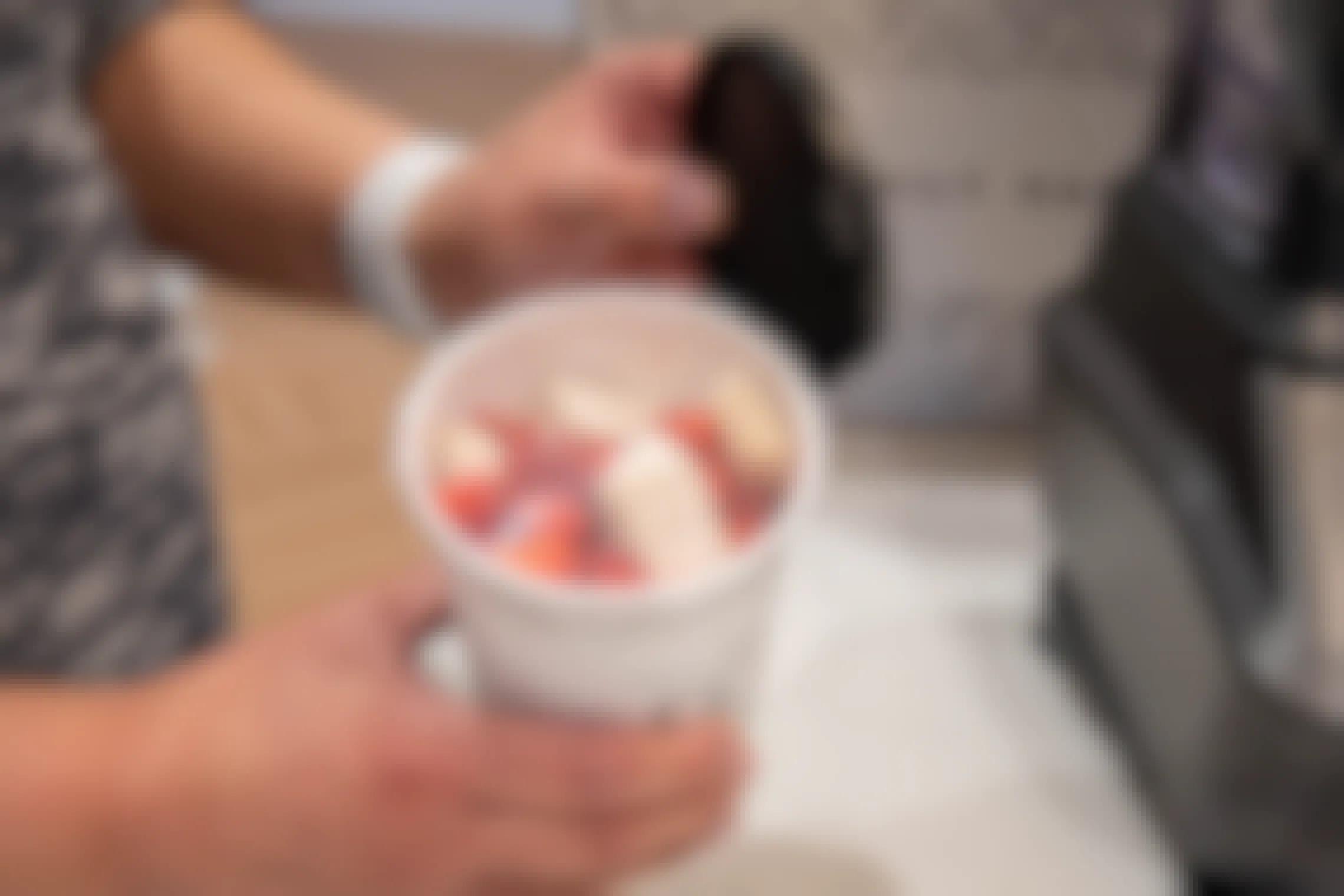 The inside of a smoothie cup showing strawberries and other fruit in front of a A woman pulling out the cups of food from a Daily Harvest meal kit box.