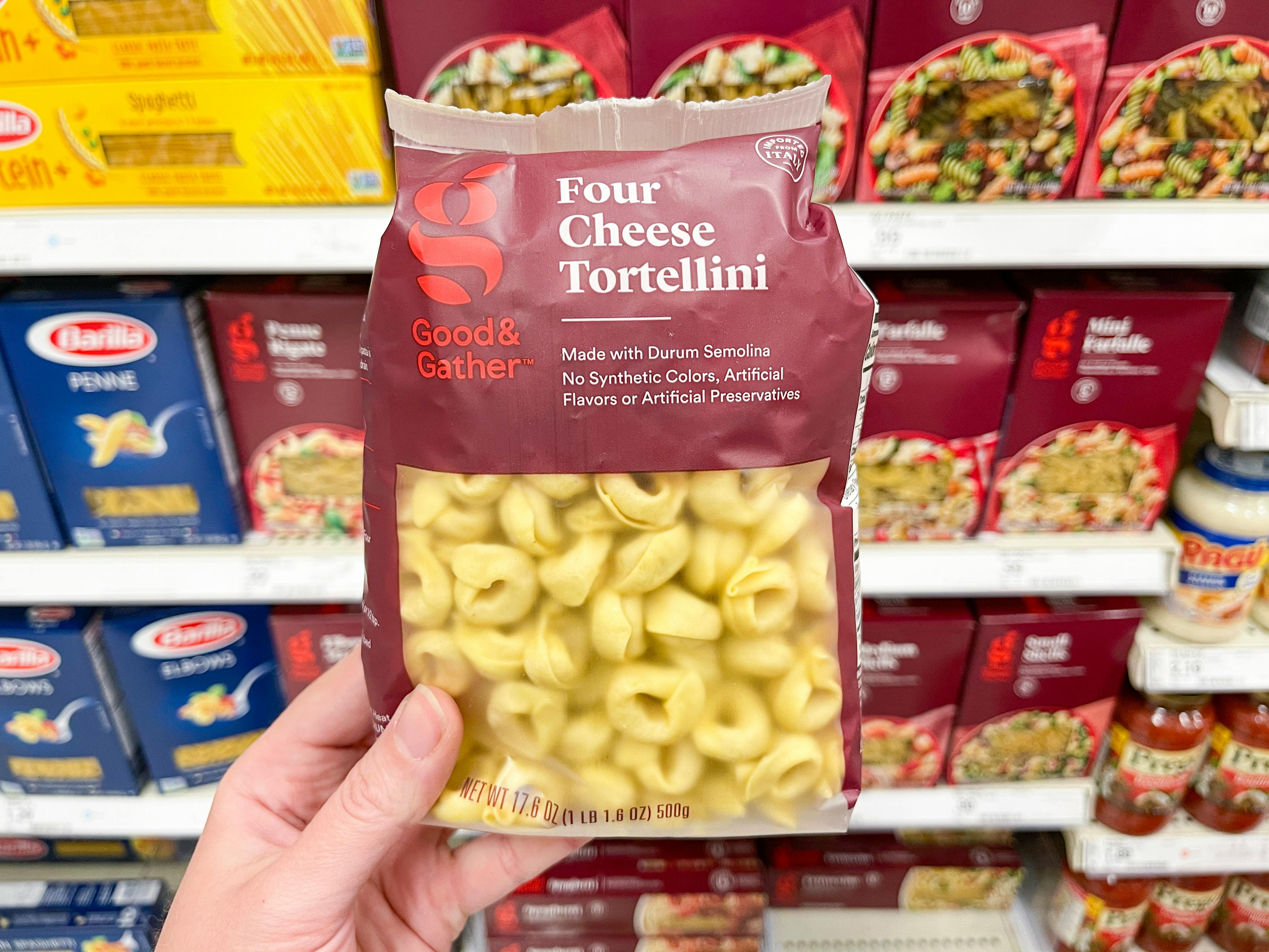 A person's hand holding a package of Good & Gather four-cheese tortellini in front of a shelf in the pasta aisle inside Target.