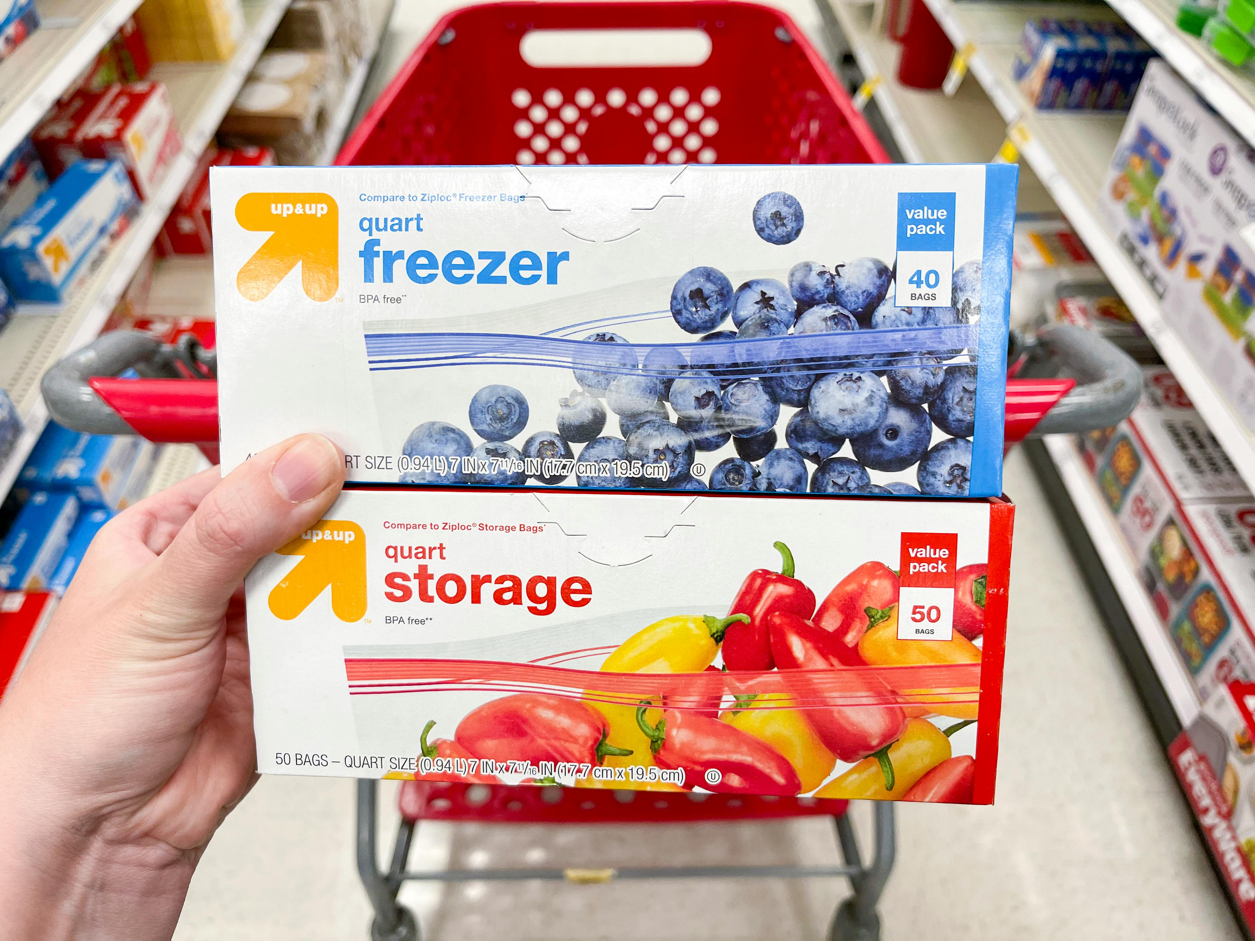 A person's hand holding two boxes of Up&Up food storage bags in front of a shopping cart at Target.