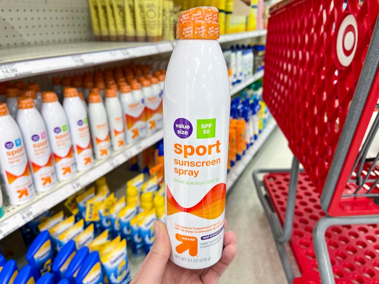 A person's hand holding a bottle of Up&UP sport sunscreen spray in front of a shopping cart in the sun care aisle at Target.