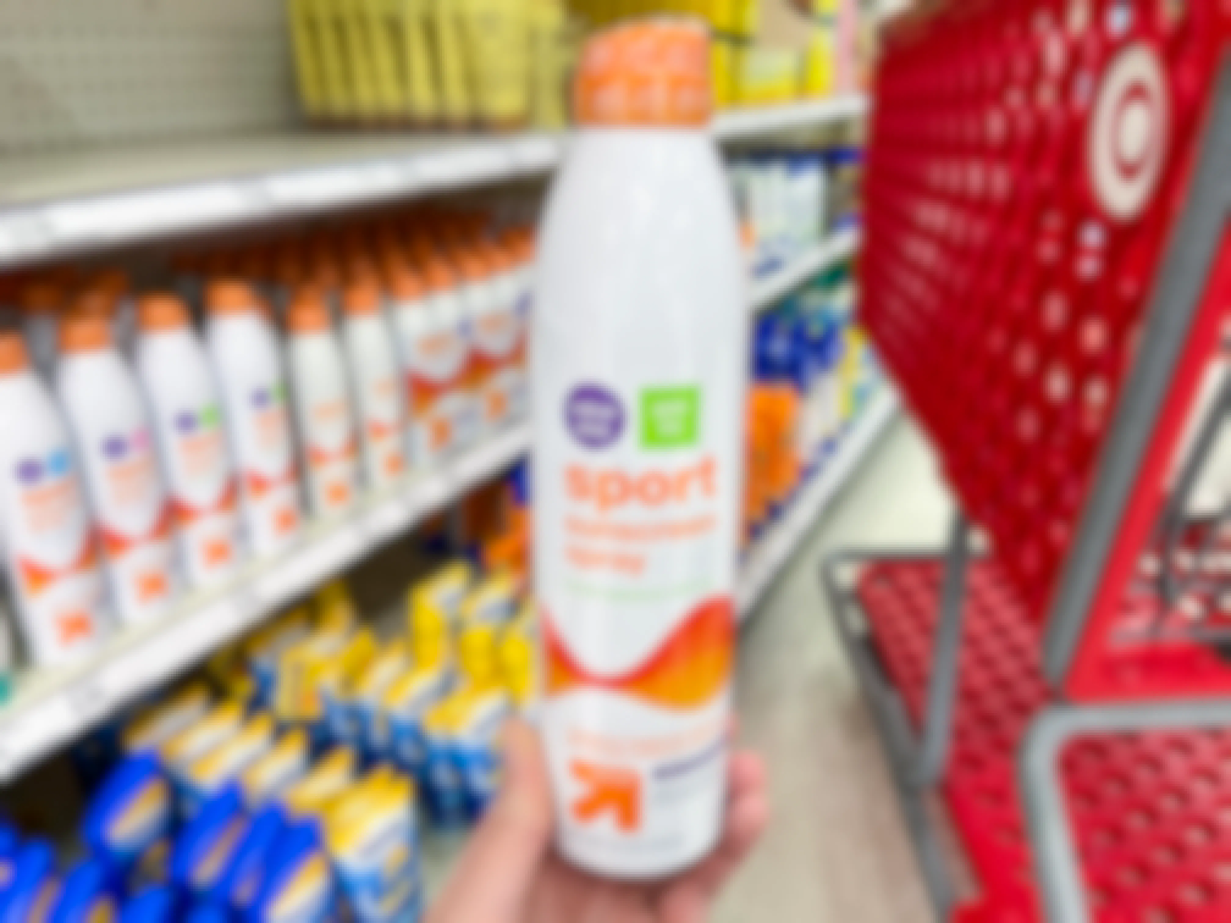 A person's hand holding a bottle of Up&UP sport sunscreen spray in front of a shopping cart in the sun care aisle at Target.