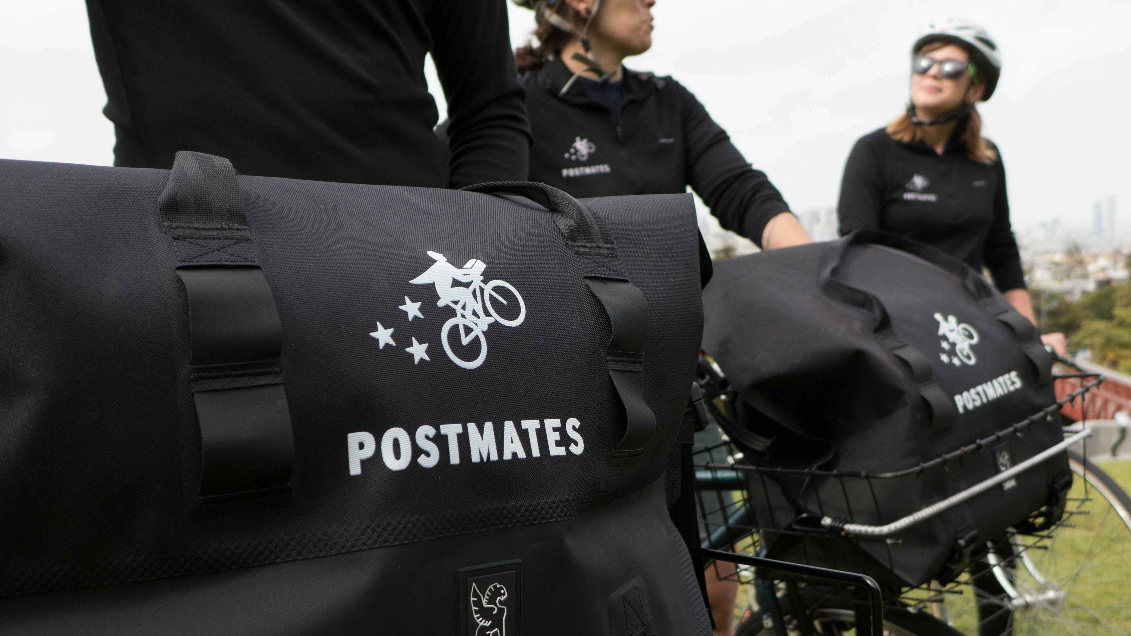 Three people on bicycles with Postmates food delivery bags in the bicycles' baskets.