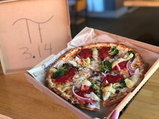 A Blaze pizza being held up inside of a box with no top. The top is displayed in the background with the Pi symbol and