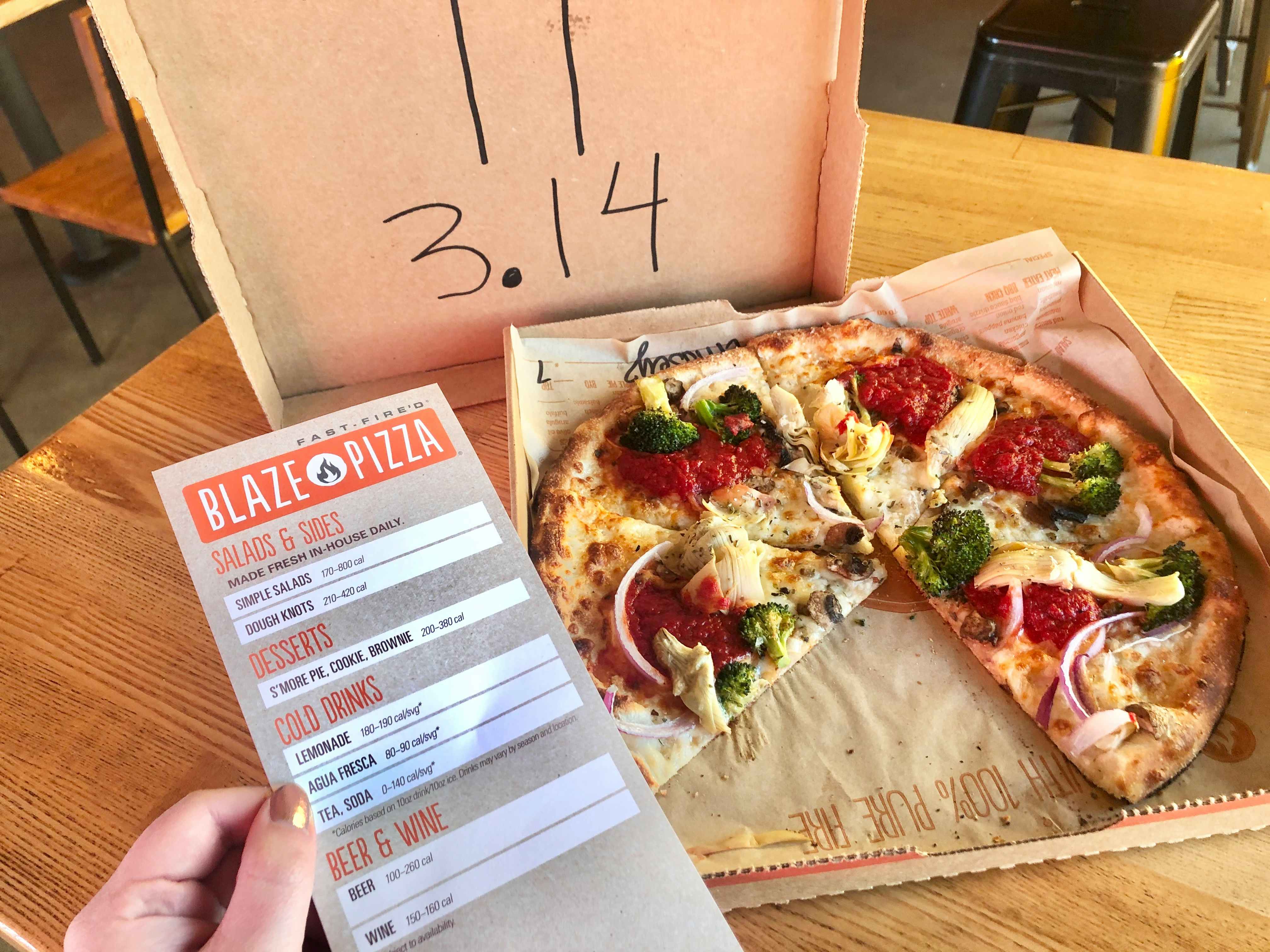 A person's hand holding the Blaze Pizza menu up in front of a pizza sitting on a table in a box with no top. The top is displayed in the background with the Pi symbol and 