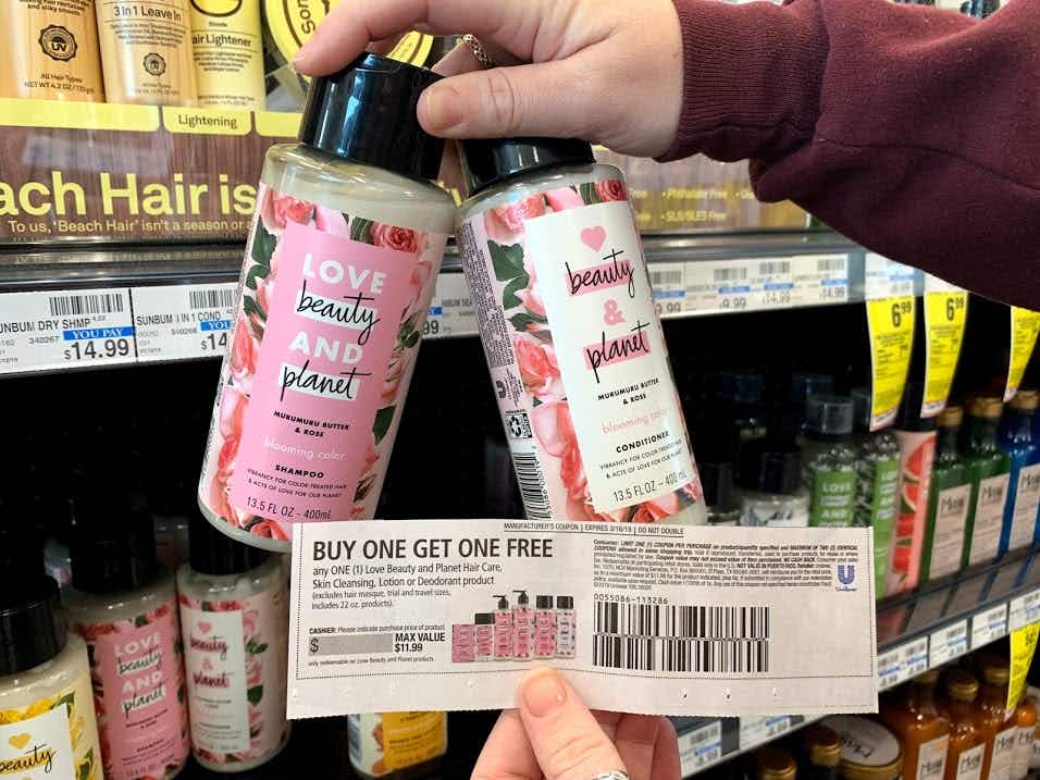 A person is standing in front of store shelves with shampoo and conditioner products while holding two bottles of shampoo with a buy one get one free coupon in front of them.