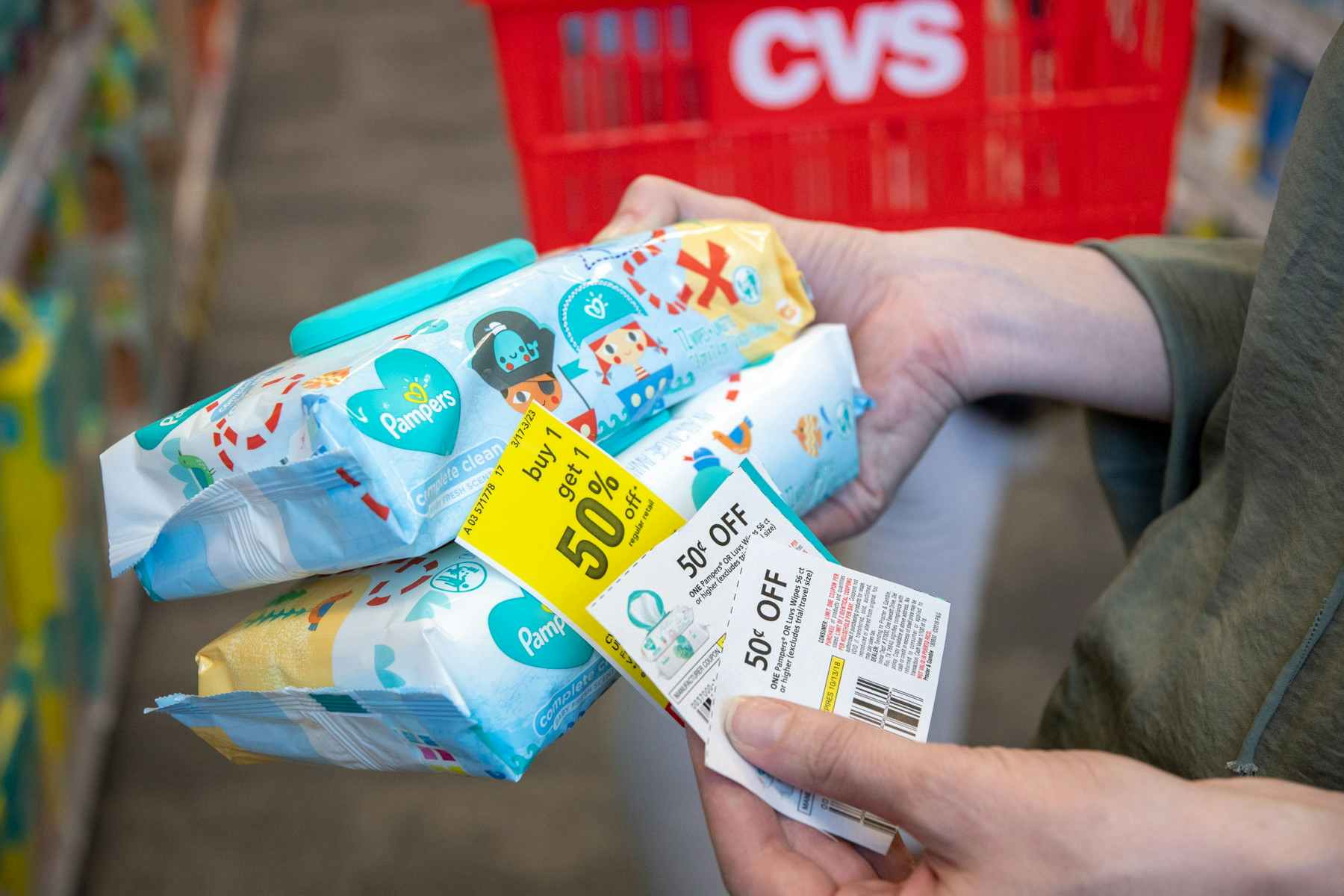 A person's hands holding two packs of Pampers baby wipes, two coupons, and a deal tag for Buy 1 Get 1 50% off in an aisle at CVS.