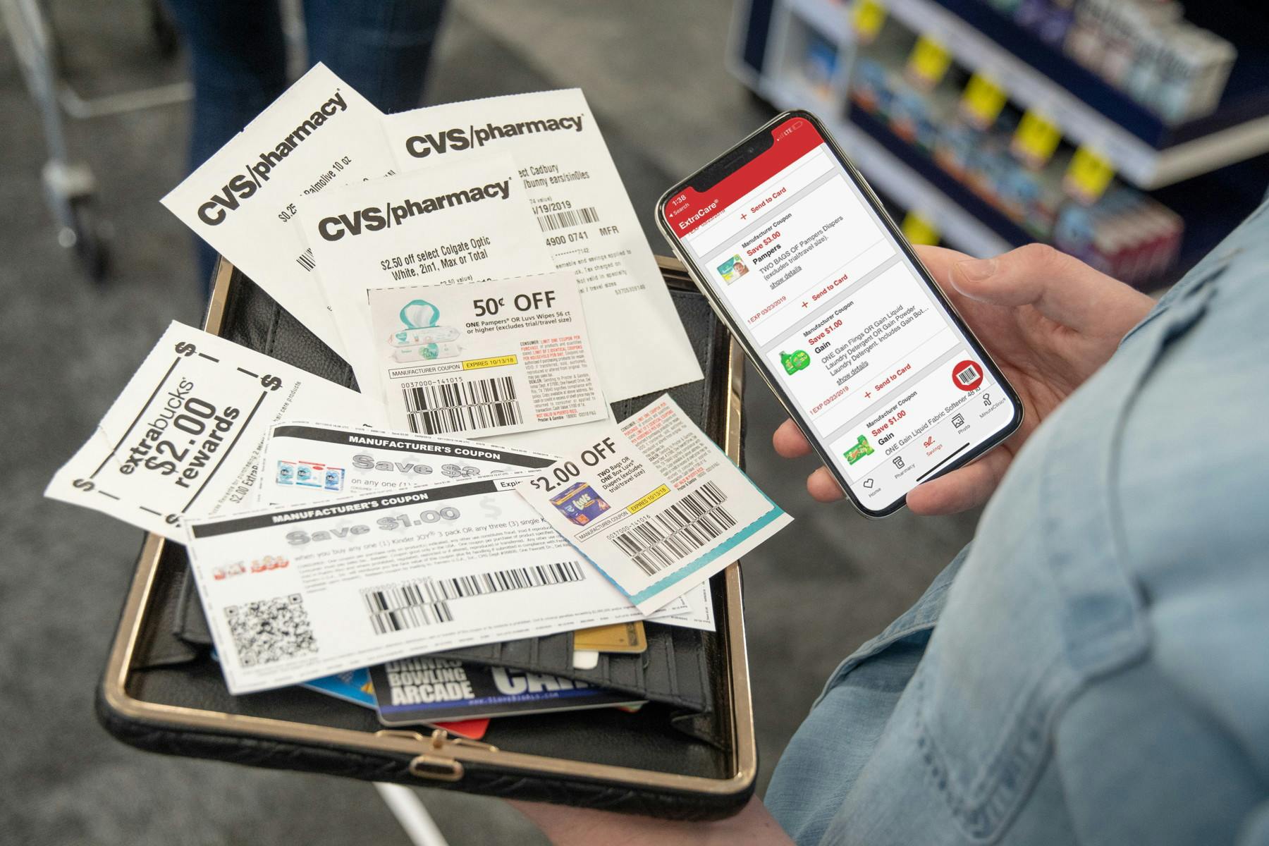 A person holding a cellphone displaying the CVS app next to a stack of coupons sitting on a wallet in the person's other hand.