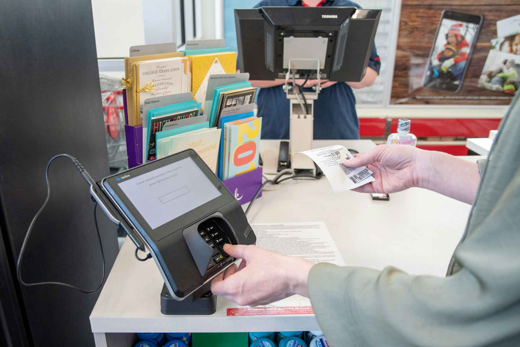A person entering numbers on a keypad at a register while holding a coupon in their other hand while a cashier stands at the register.