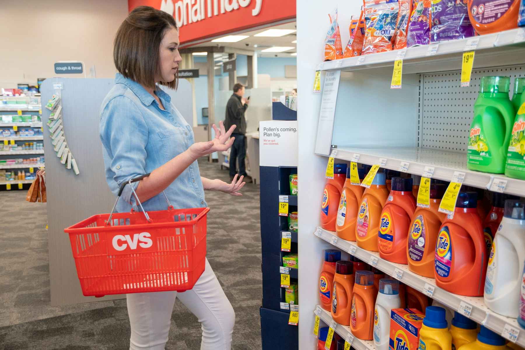 A woman standing a store with a CVS basket on her right arm while looking at shelves with laundry detergent, with one shelf empty.