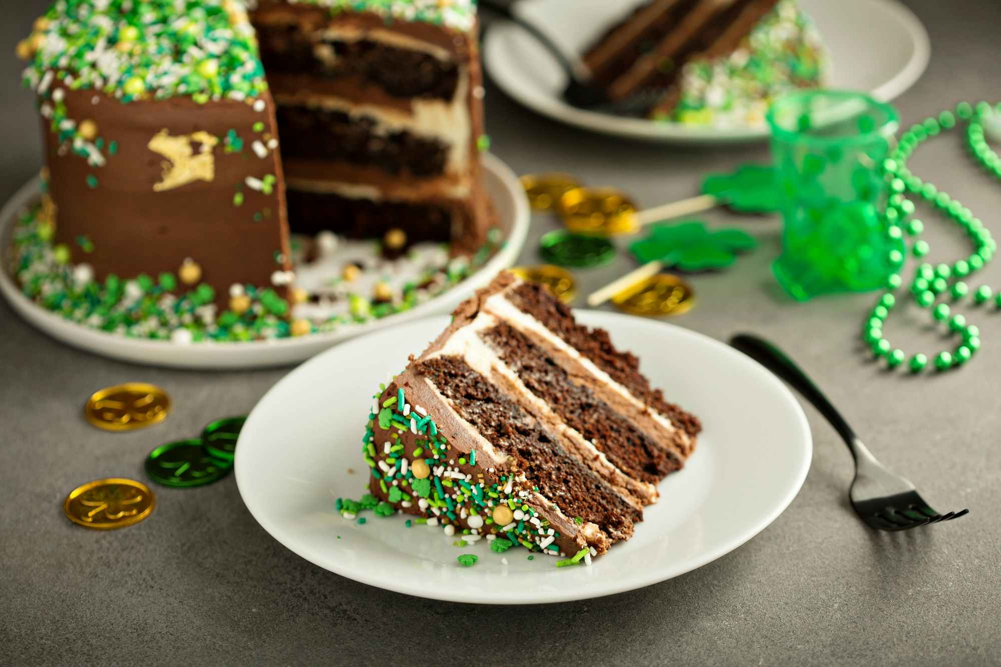 A piece of chocolate cake with green sprinkles on a plate next to the rest of the cake and some St. Patricks Day themed items
