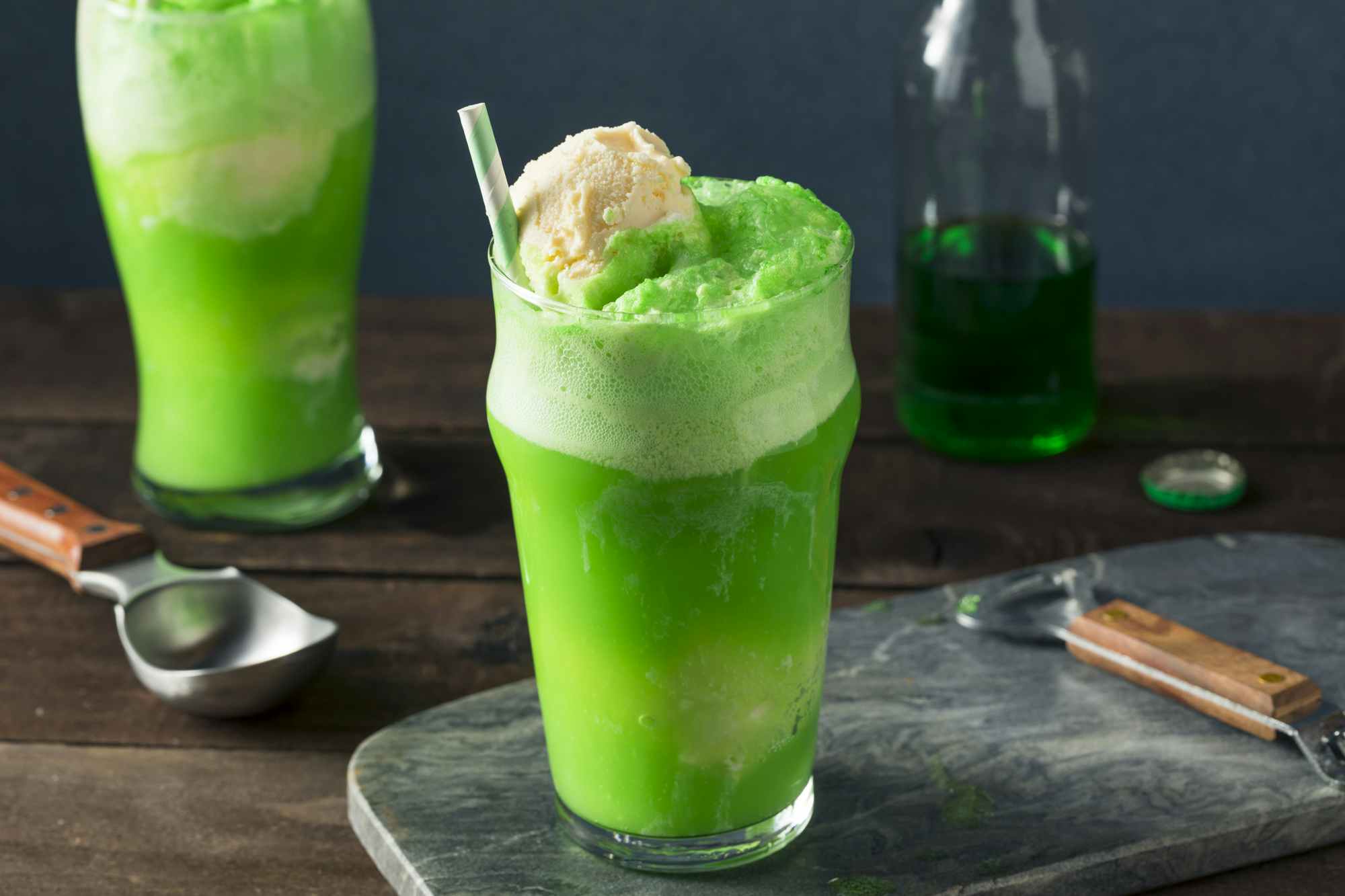 Green soda floats on a table with an ice cream scoop, a soda bottle, bottle opener, and bottle cap