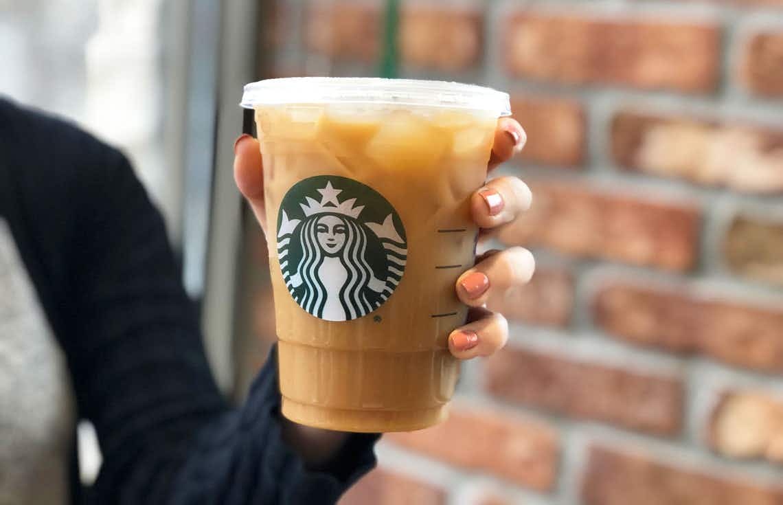 A person holding up a Starbucks iced coffee in front of a brick wall.
