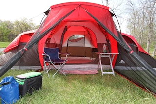 A large red Ozark tent set up with a table and chair and some coolers in a field.