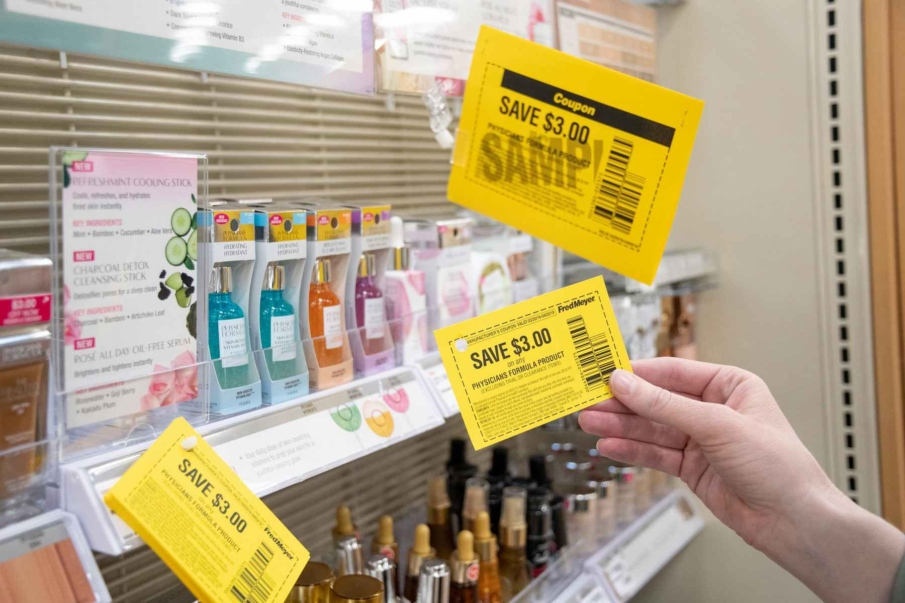 A person's hand holding up a coupon for $3 off next to a shelf with fragrances and more coupons hanging from a hook.