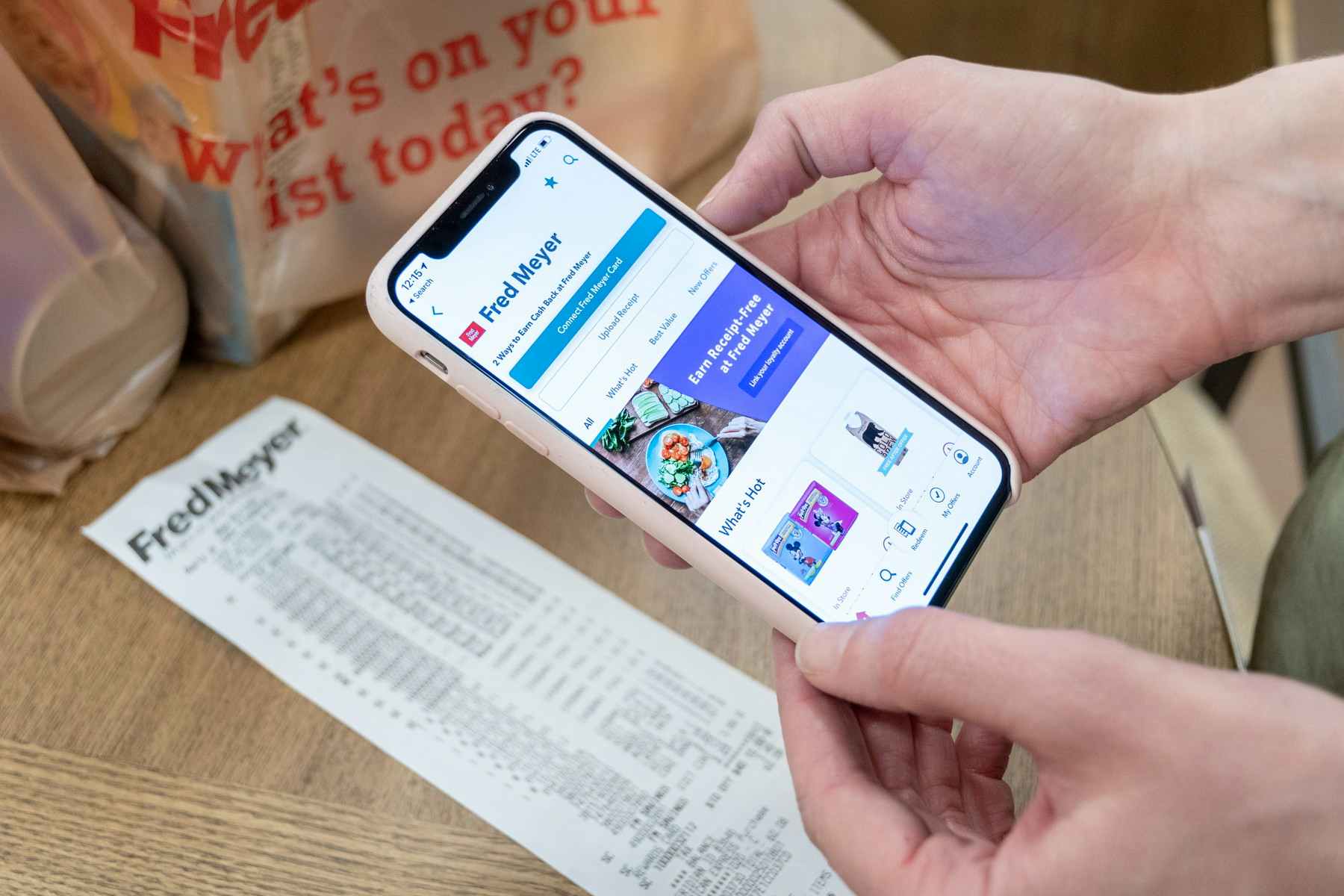 A person's hands holding an iPhone with the Ibotta mobile app open about to scan a receipt that is on a table next to two bags of groceries.