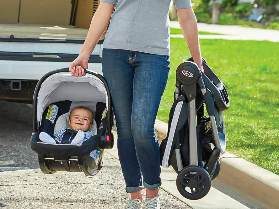 A woman carrying a Greco car seat and foldable stroller outside.