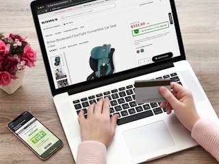 A person shopping on the Kohl's website for car seats with their phone displaying kohls cash on the table next to the laptop.