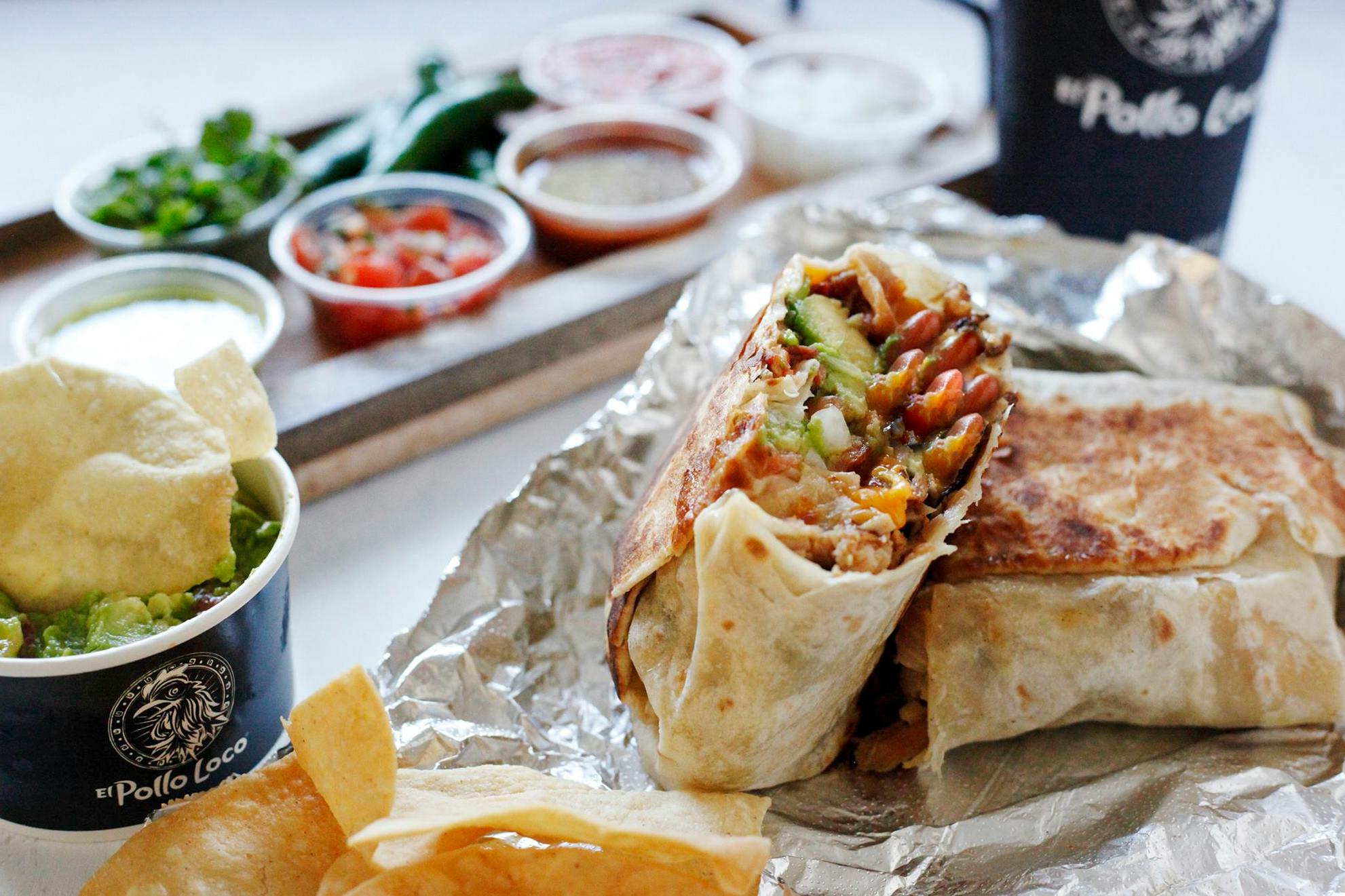 A burrito from El Pollo Loco, cut in half, sitting on foil next to a tray of dipping cups with salsa, a cup of guacamole with a chip in it, and a drink cup.