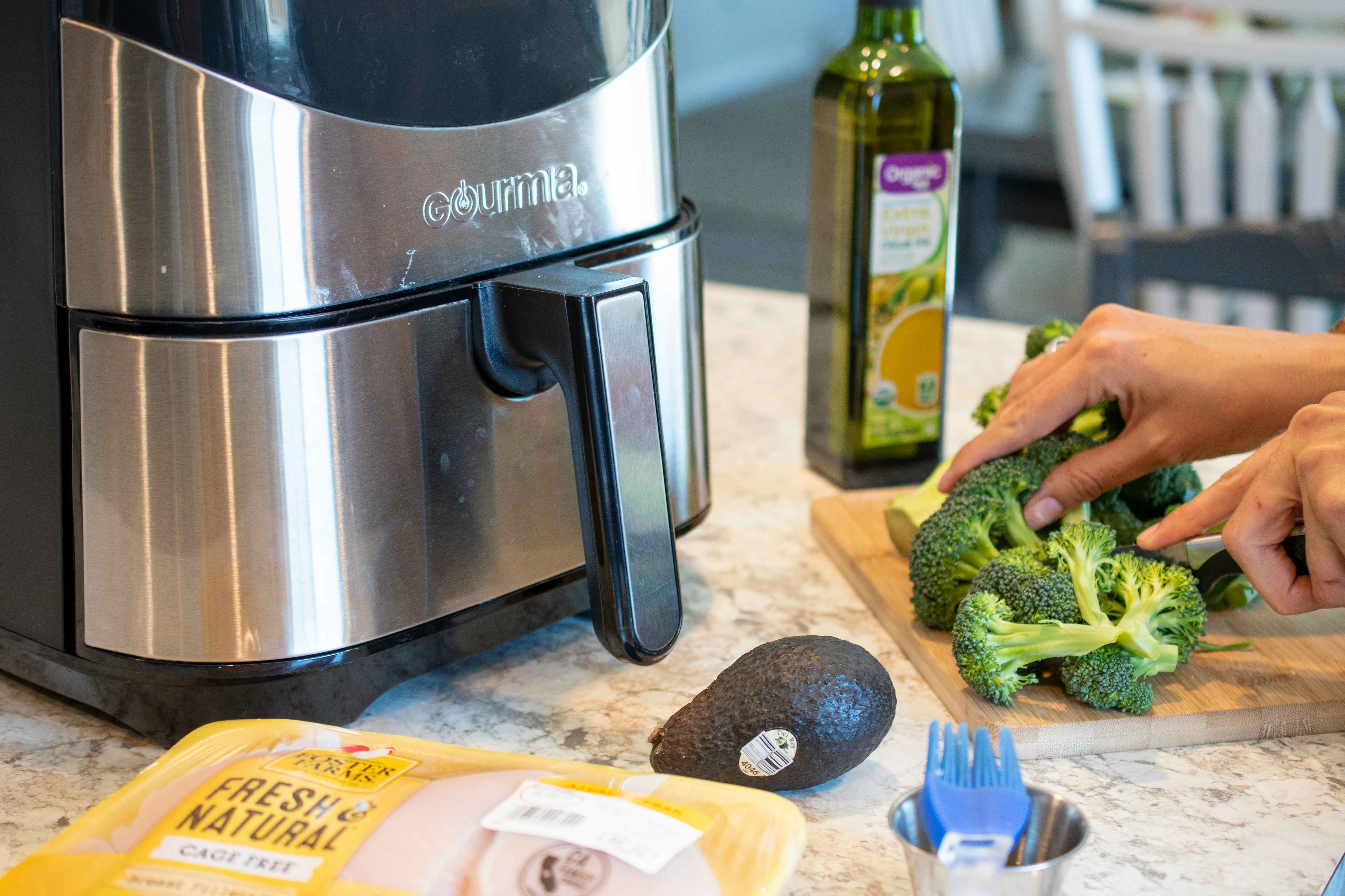 A person cutting broccoli next to other ingredients and an air fryer