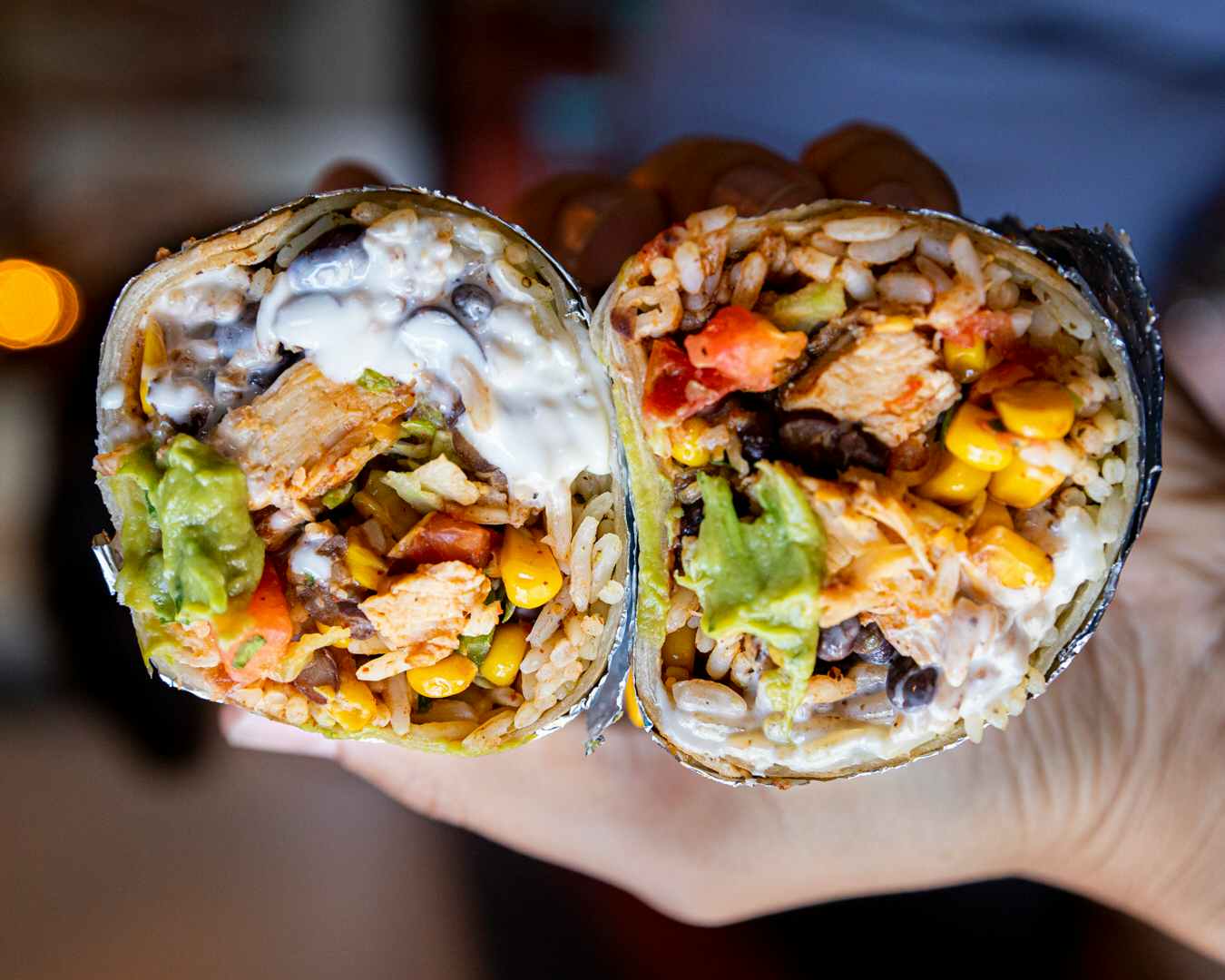A person's hand holding up two halves of a burrito, showing the inside. The burrito is filled with meat, cheese, beans, rice, vegetables, sour cream, and guacamole.
