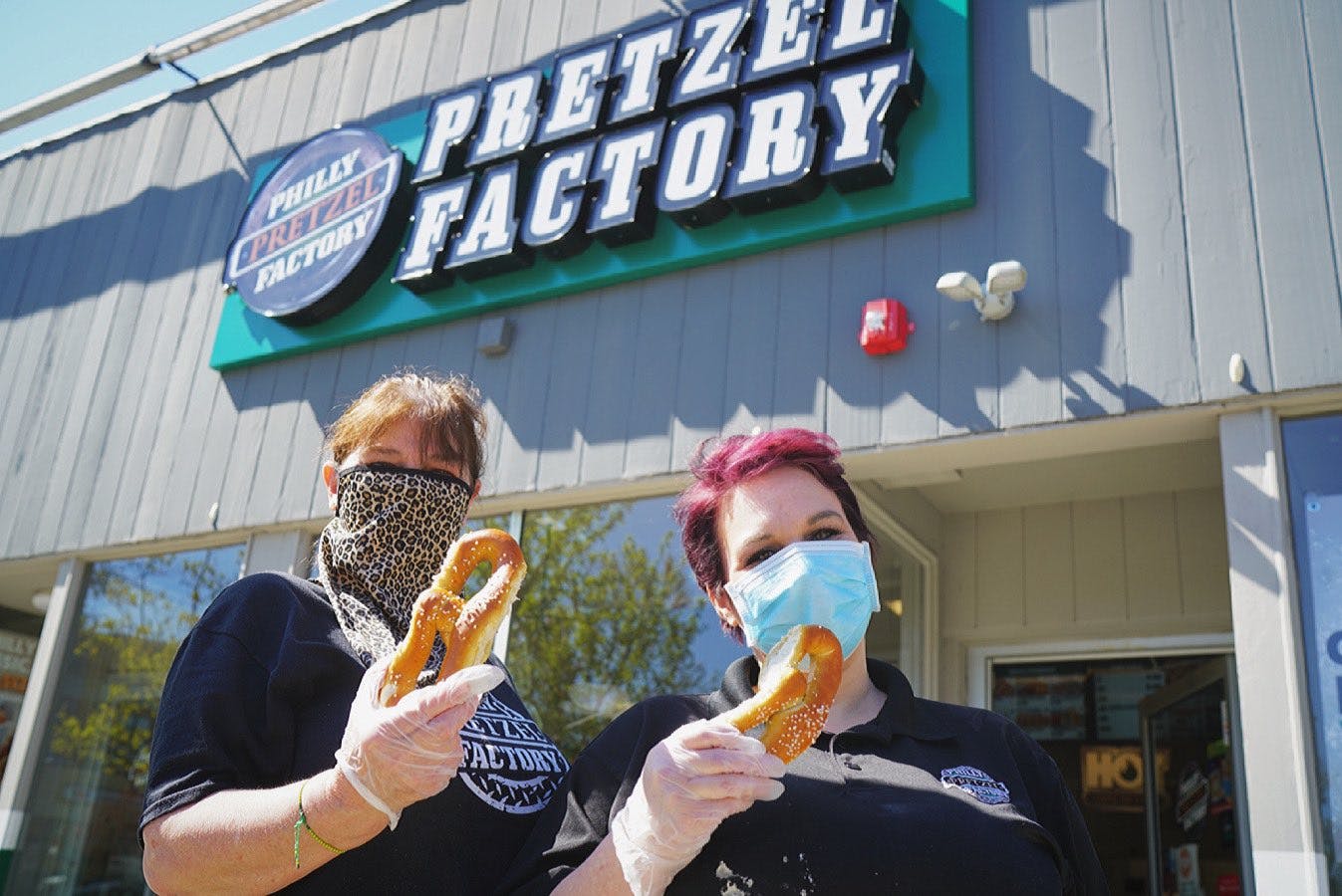 Two people in masks standing outside the Philly Pretzel Factor, each holding up a soft pretzel.