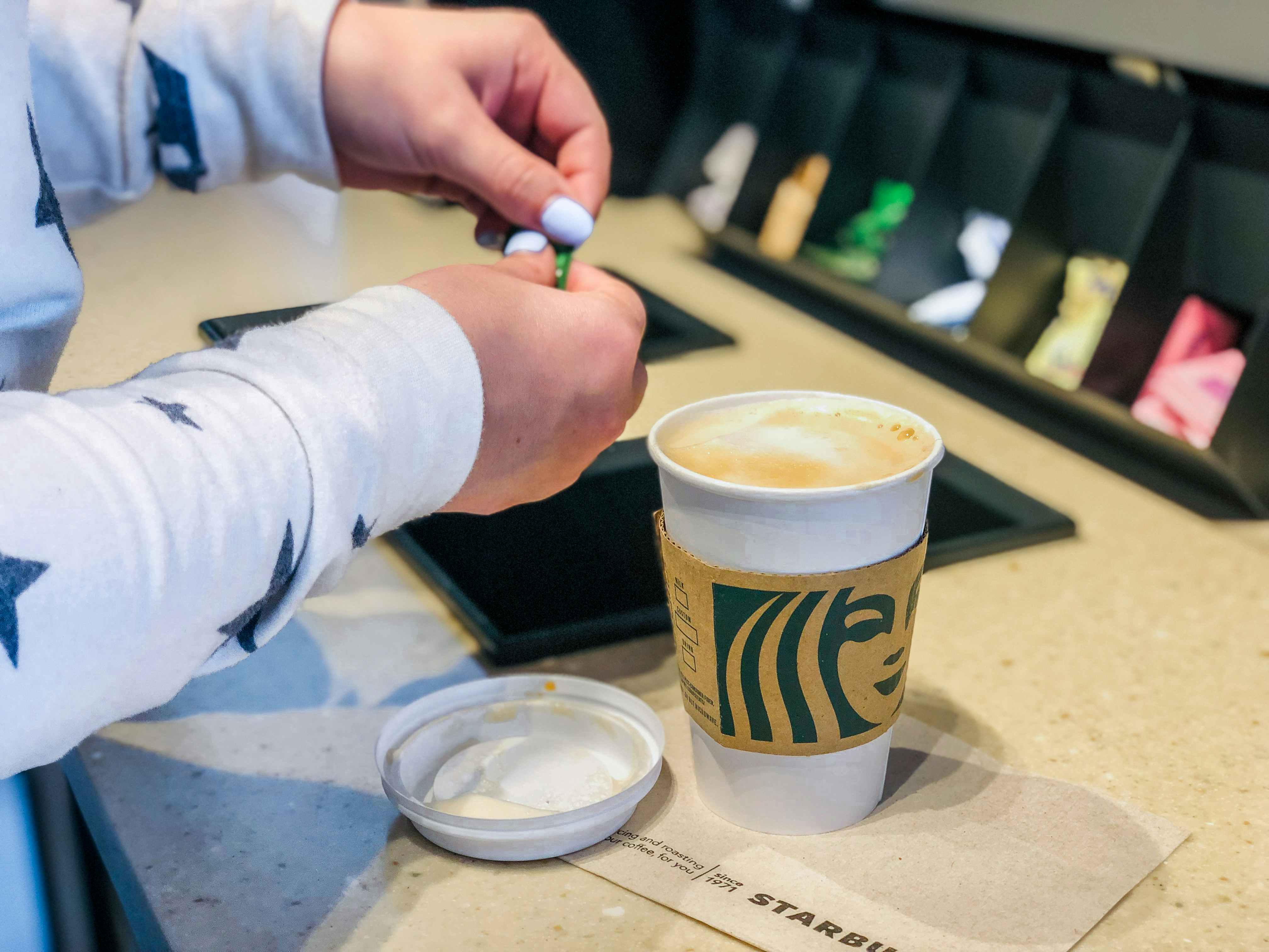 A person opening a pack of sugar into their coffee sitting on the counter with the lid off at Starbucks.