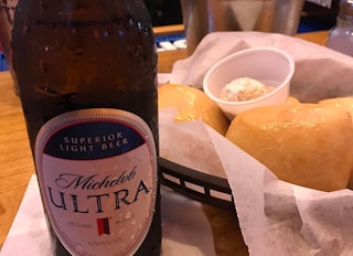 A close-up of a bottle of Michelob Ultra beer next to a basket of bread and butter on the table at Texas Roadhouse.