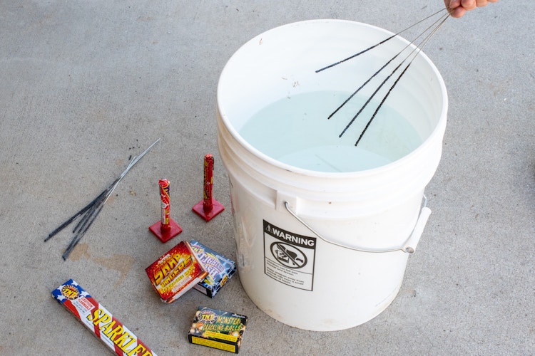 Someone extinguishing some sparklers in a bucket of water on the ground next to more sparklers and other small fireworks.