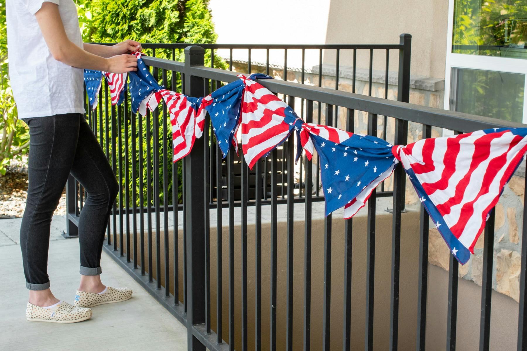 A person affixing a 4th of July bunting to a metal fence that was made by tying American flag bandanas together. 