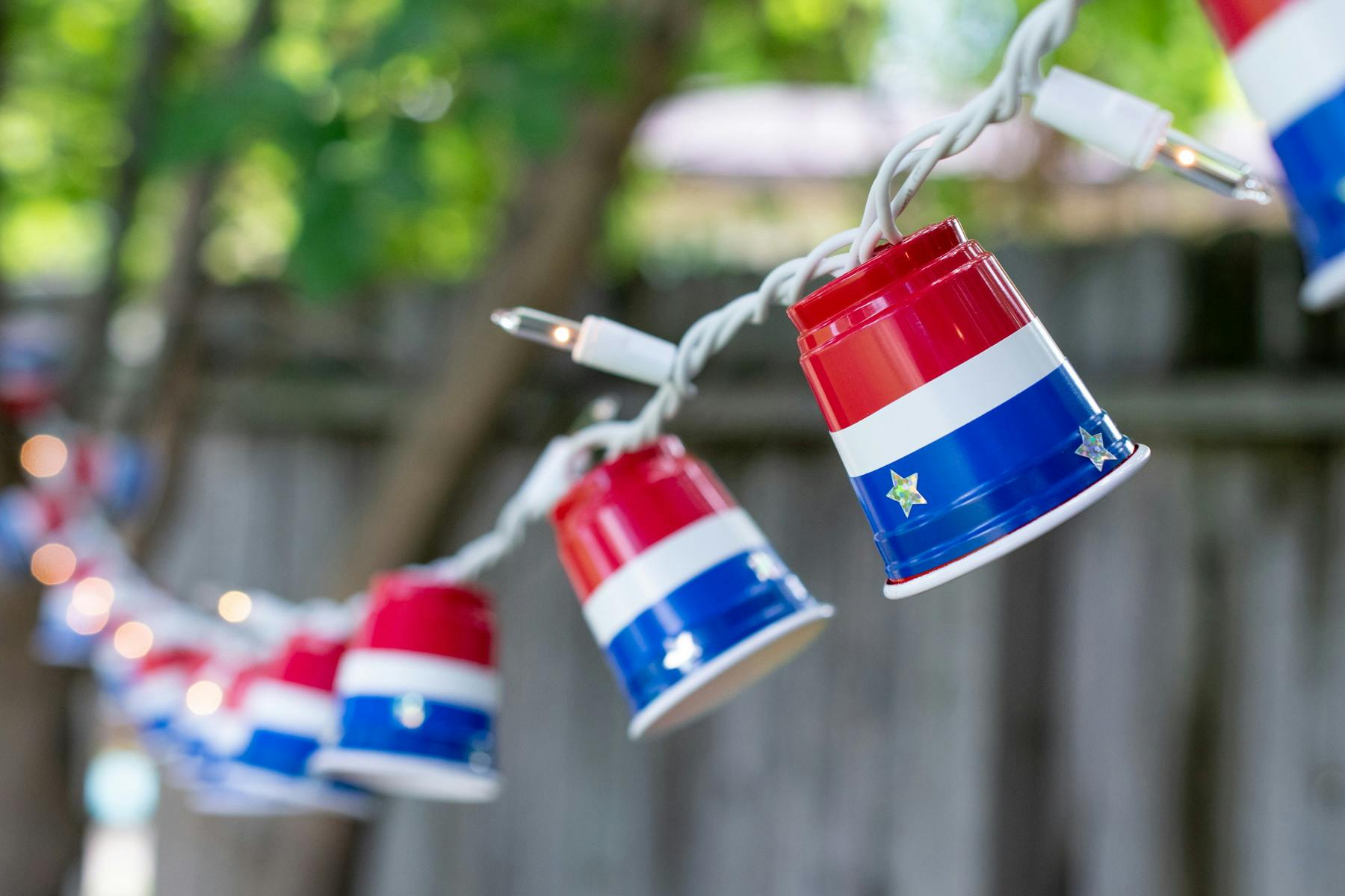 Red plastic shot glass cups affixed to party lights with blue and white electrical tape and star stickers hanging across a backyard.