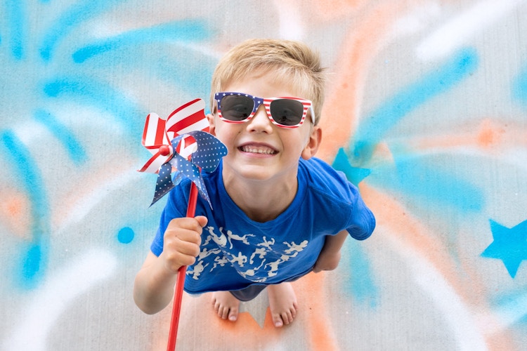 A child with American flag sunglasses and an American flag spinner standing on a 4th of July firework photo backdrop painted onto the concrete with spray chalk.