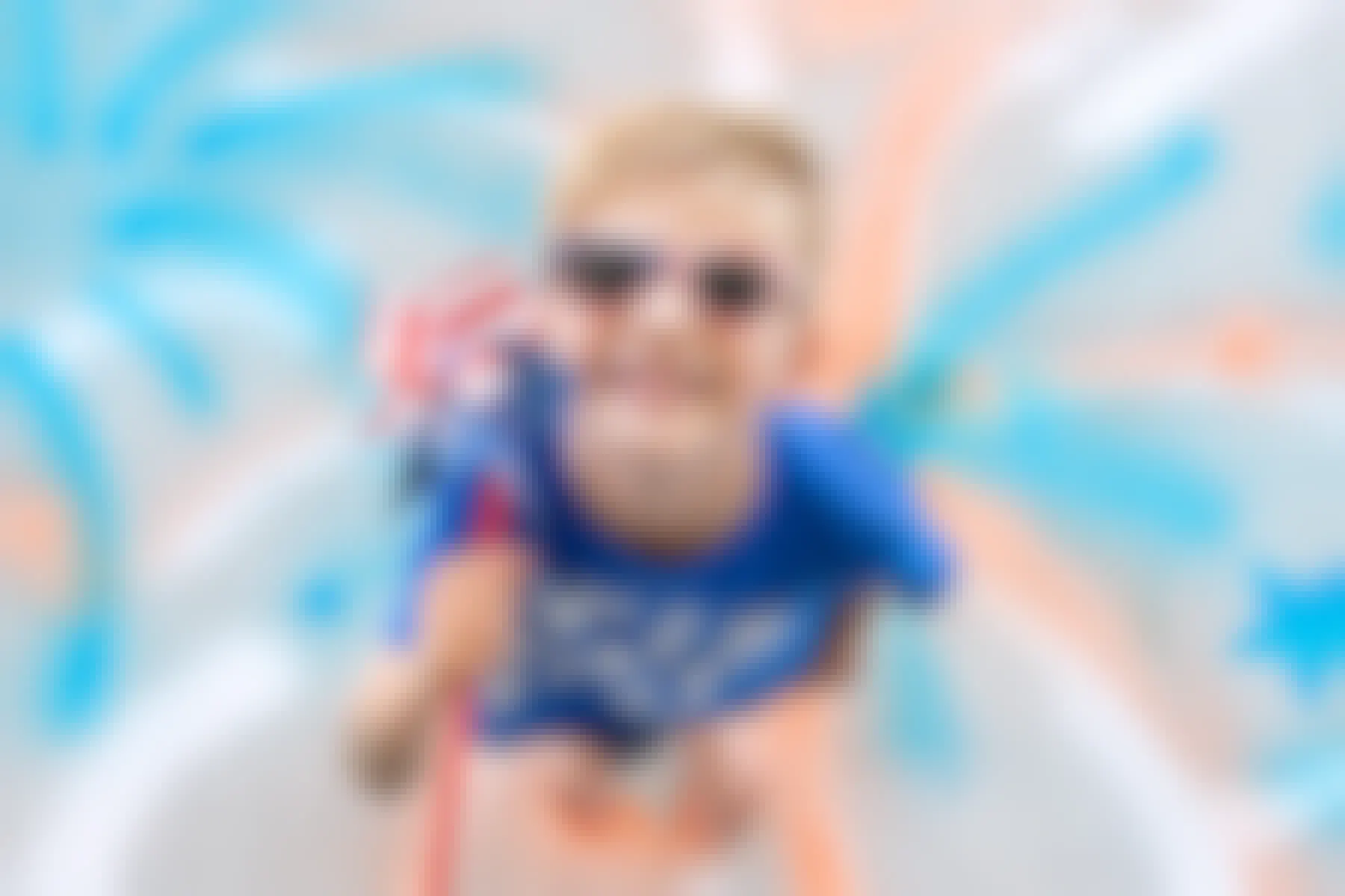 A child with American flag sunglasses and an American flag spinner standing on a 4th of July firework photo backdrop painted onto the concrete with spray chalk.