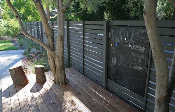 A chalkboard hanging from a backyard fence with some chalk drawings on it.