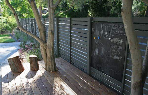 A chalkboard hanging from a backyard fence with some chalk drawings on it.