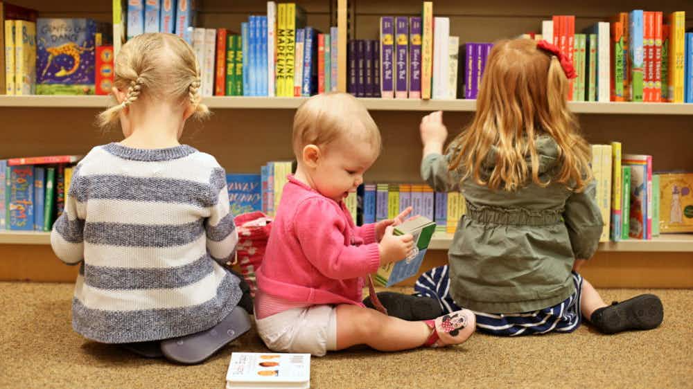 Three children sitting on the floor reading books in front of a bookcase filled with books.
