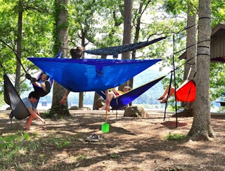 People sitting in and standing around some camping hammocks strung up between trees near a lake.