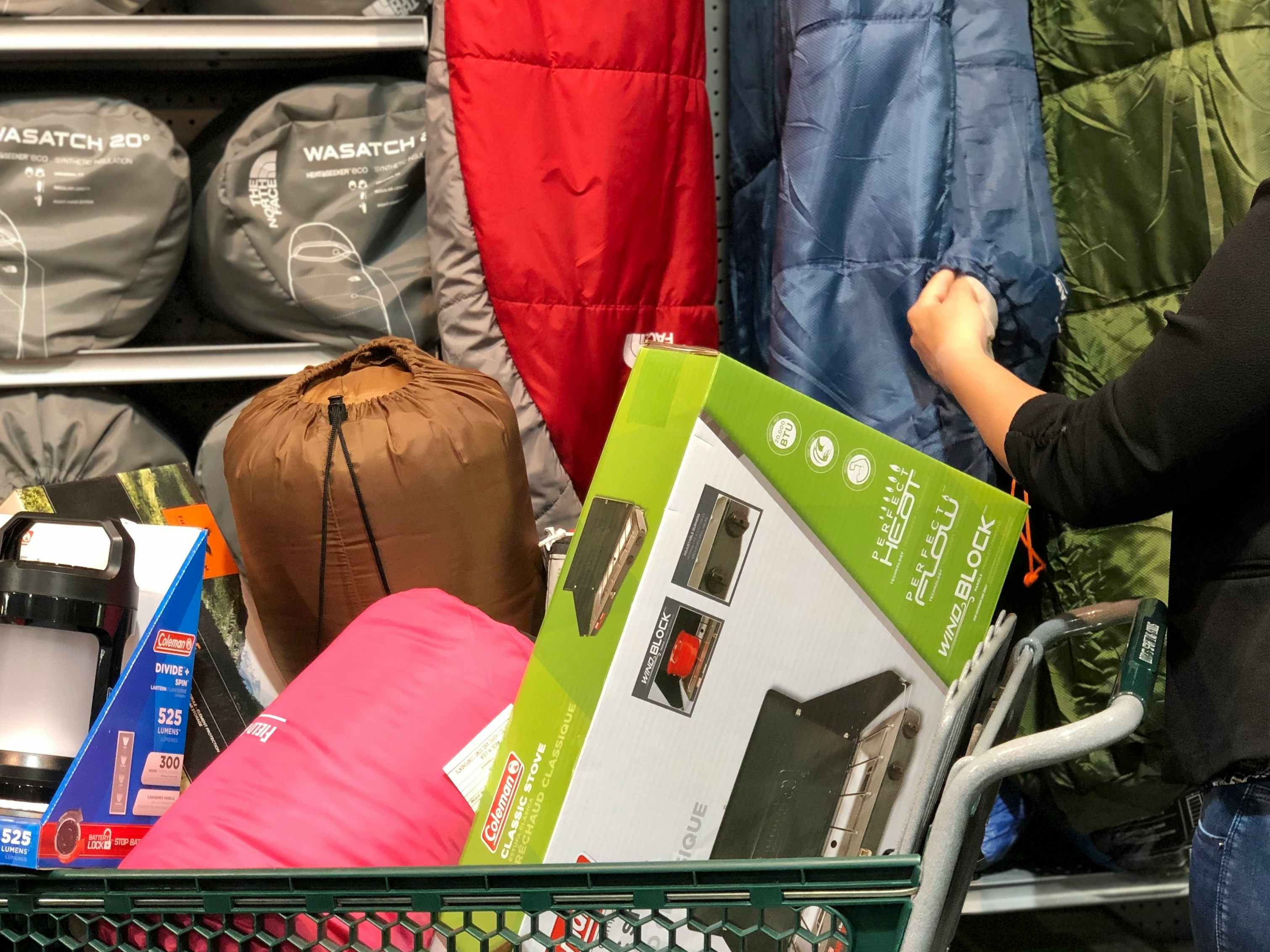 A person looking through sleeping bags next to a shopping cart filled with camping gear including an air mattress and sleeping bags.
