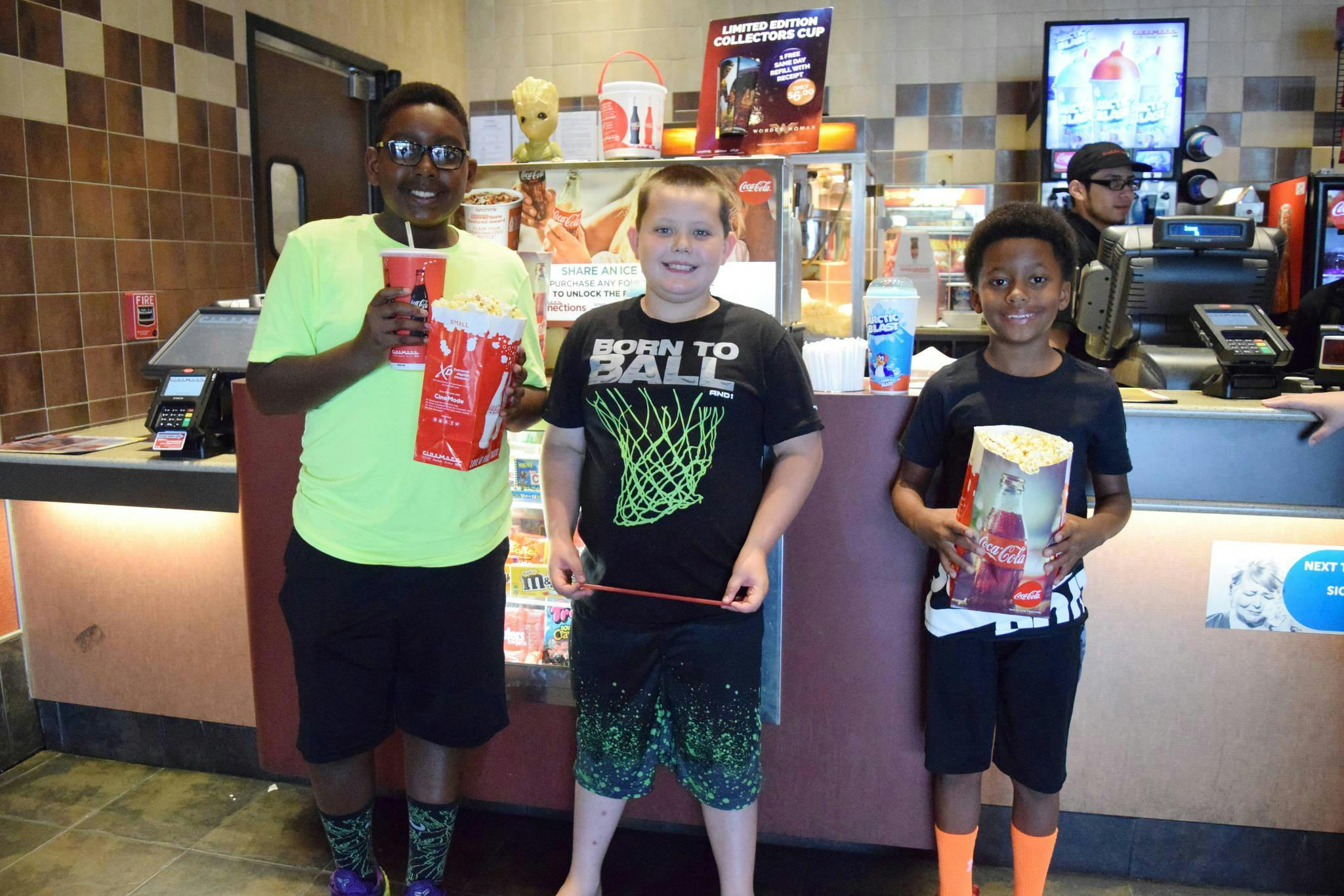 Three kids standing in front of a movie theater concession counter, holding bags of popcorn and drinks.