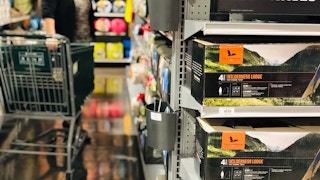 A person pulling a shopping cart down the camping gear aisle at Dick's Sporting Goods, with two Field and Stream tent boxes sitting on the shelf in the foreground.