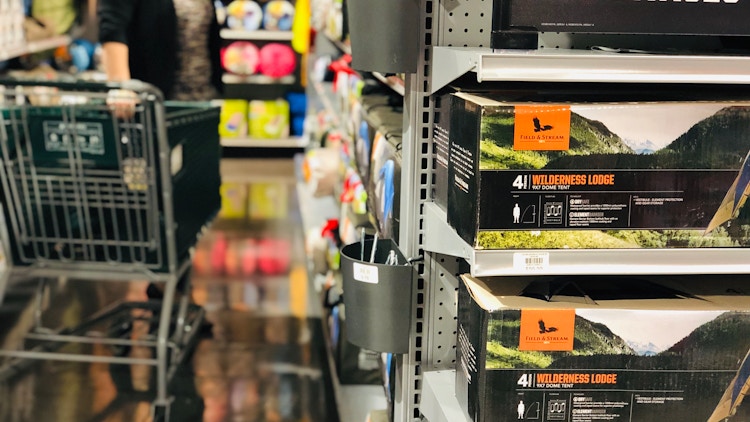 A person pulling a shopping cart down the camping gear aisle at Dick's Sporting Goods, with two Field and Stream tent boxes sitting on the shelf in the foreground. 