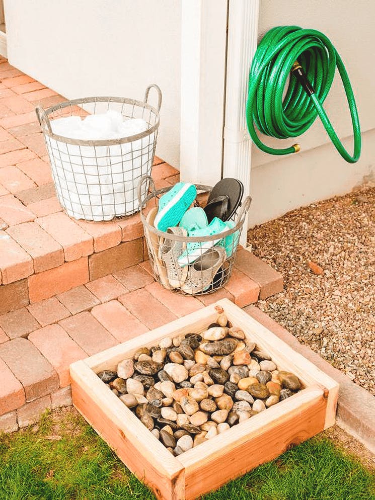 A foot washing station off of the back entrance of a home with a garden hose wound up, a box of rocks, a basket of shoes, and a basket of towels.
