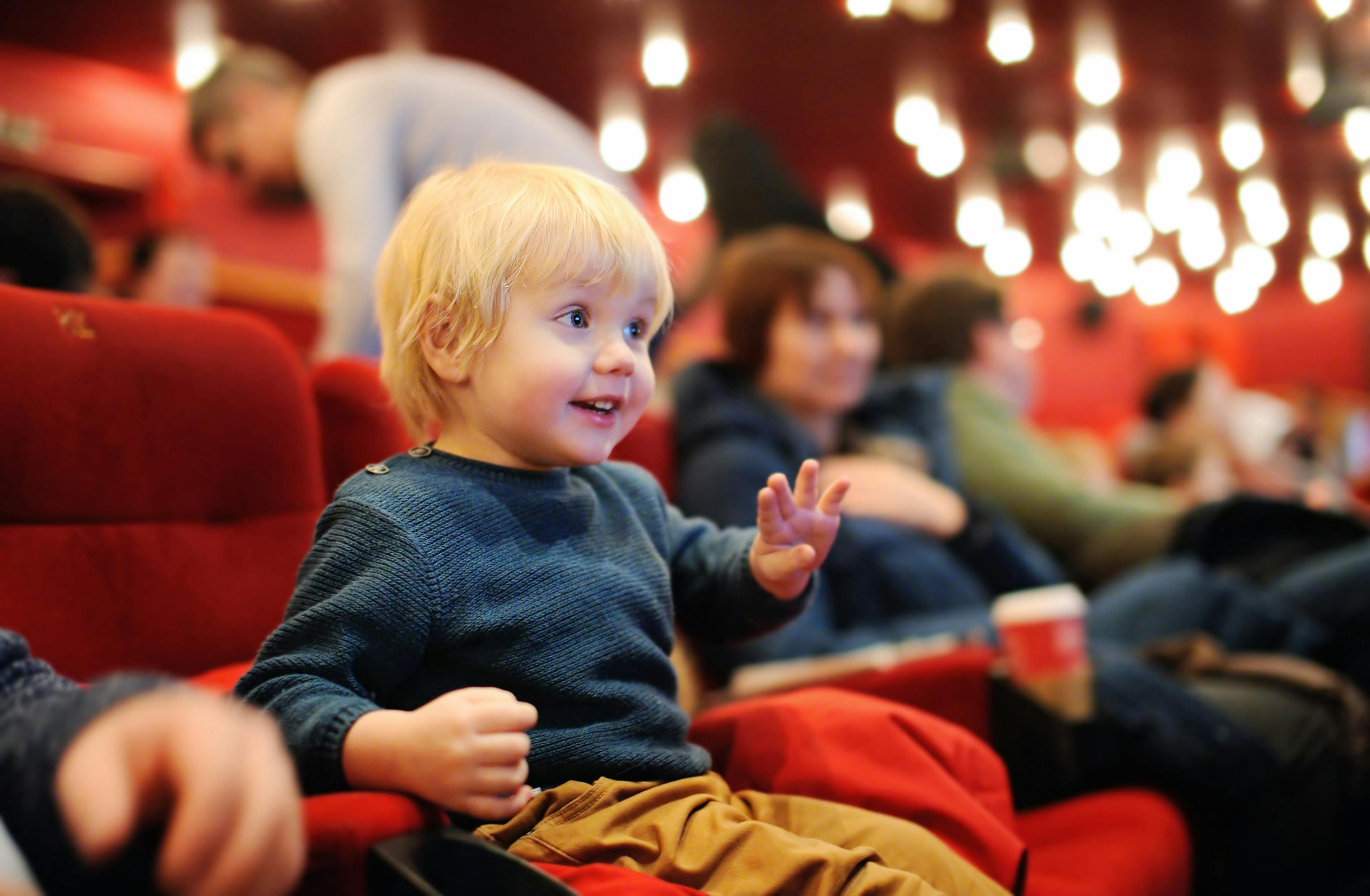A toddler sitting in a movie theater seat and looking excitedly toward the screen.