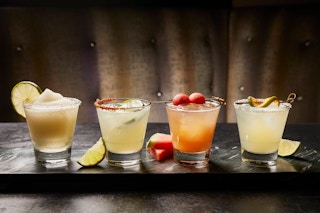 Four cocktail drinks in glasses sitting on a table with fruit garnishes.