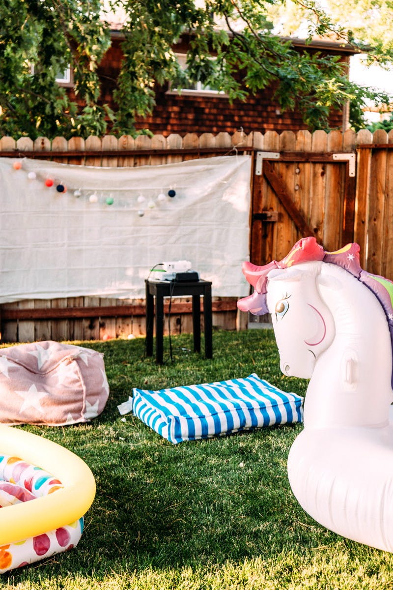 A backyard set up for a movie night with a white sheet hung on the fence, a projector set up, and various beanbags and pool inflatables scattered around as seating.